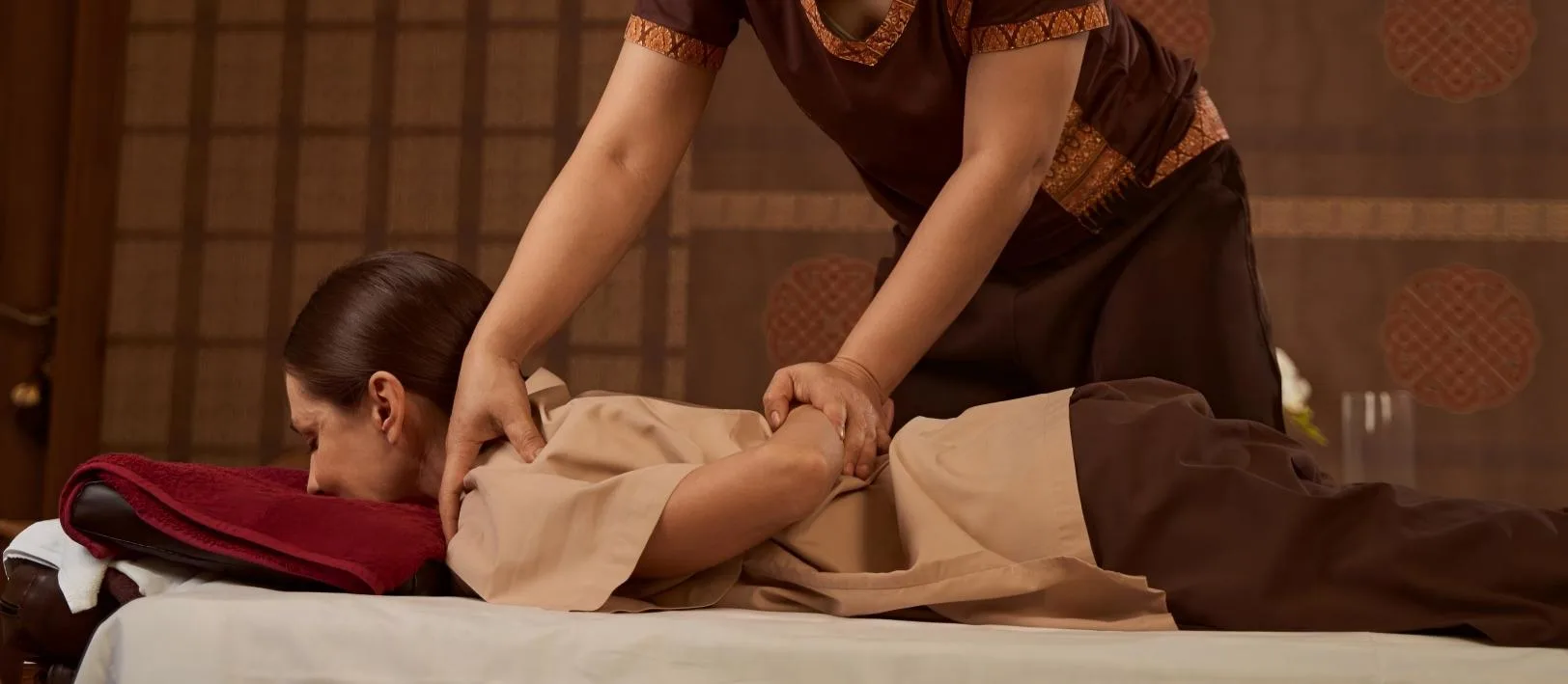 Thai massage — therapist in patterned Thai silk working a client's arm, Oriental screen backdrop